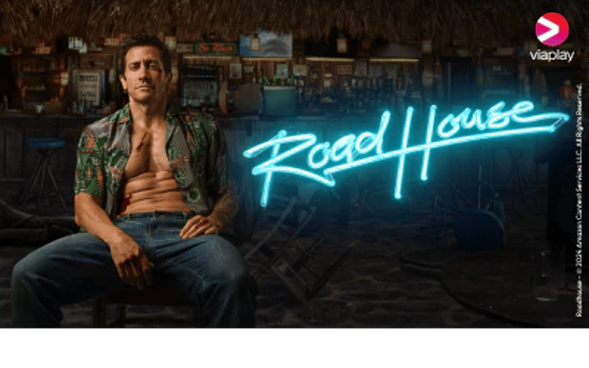 Road House på Viaplay med en mand siddende foran en bar og den neonlysende titel i baggrunden.