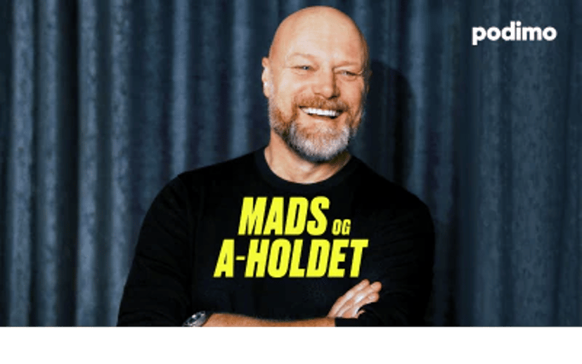 Portræt af en smilende, skaldet mand i sort trøje med teksten “Mads og A-holdet” foran en mørk gardinbaggrund.