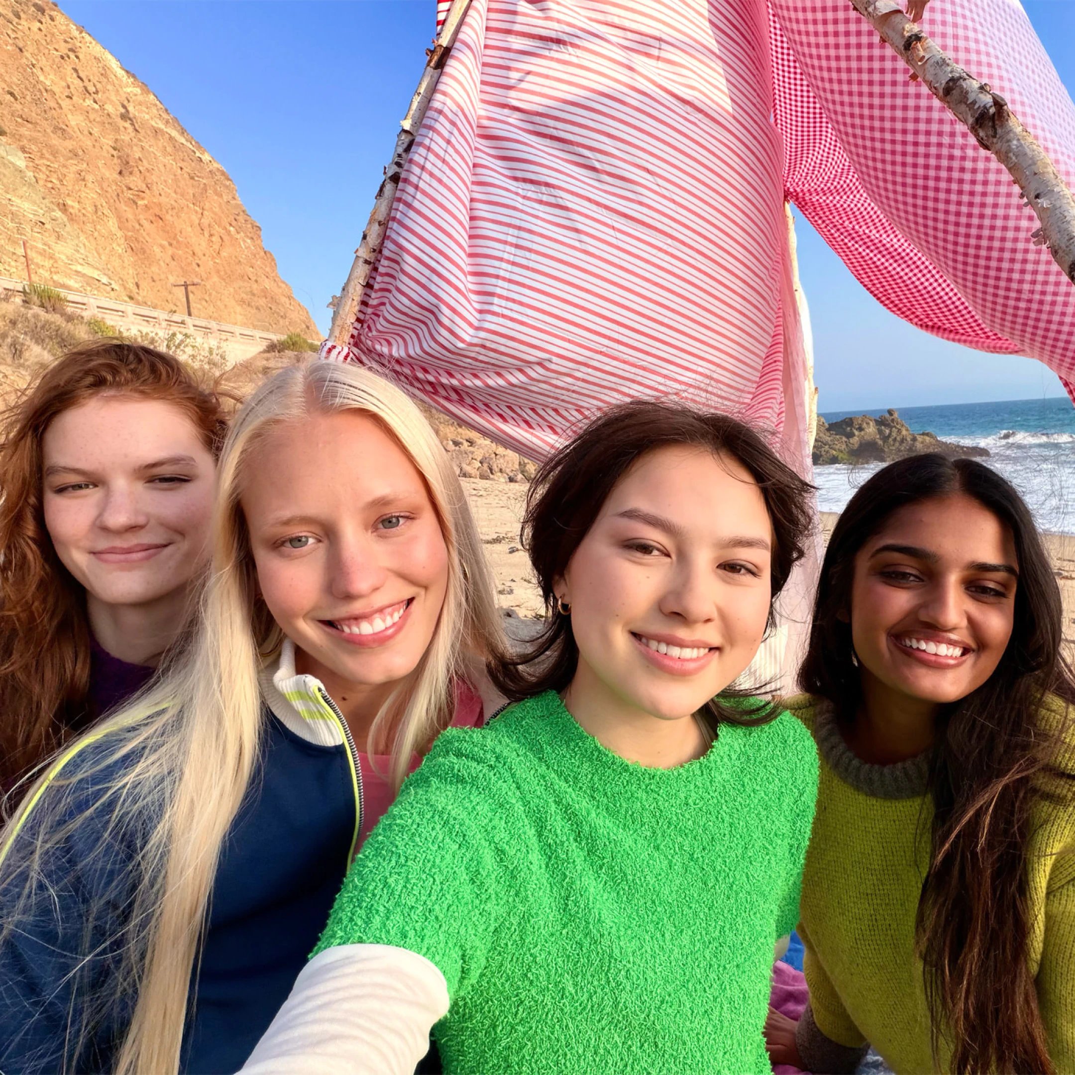 Selfie af fire smilende personer foran en strand med klipper, hav og et rødt- og hvidstribet vindsejl i baggrunden.