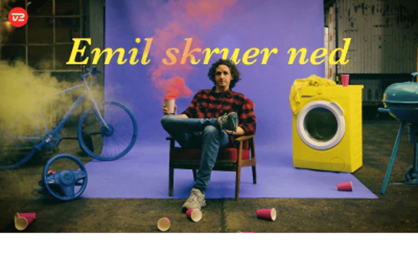 Emil sidder i en stol mellem en cykel og en gul vaskemaskine i en farverig reklameopsætning for TV 2 Play og CBB Mix.