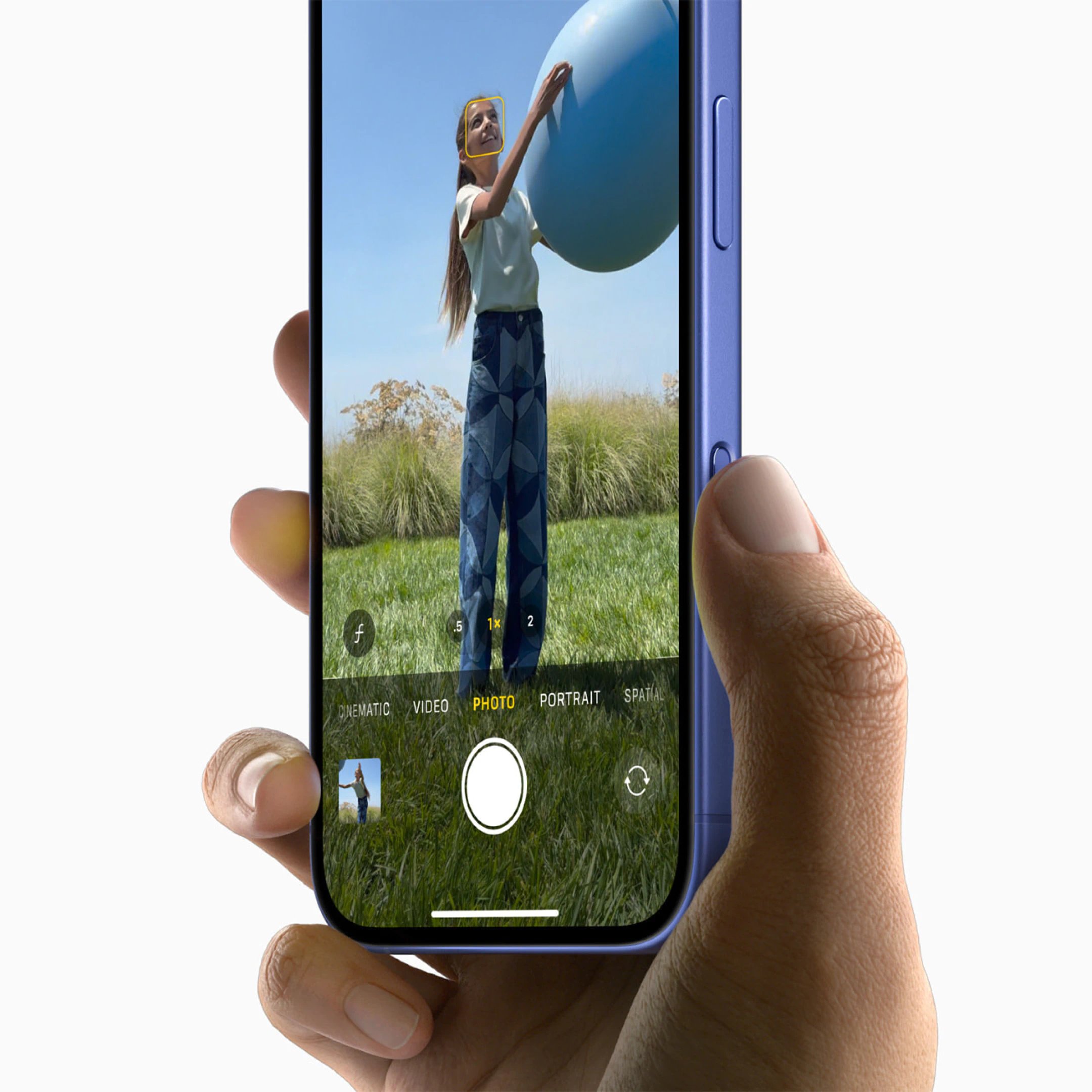 En hånd holder en blå iPhone 16, hvis kamera viser en person udendørs med en stor ballon. Kameraappen er indstillet til fototilstand med motivgenkendelse omkring ansigtet.