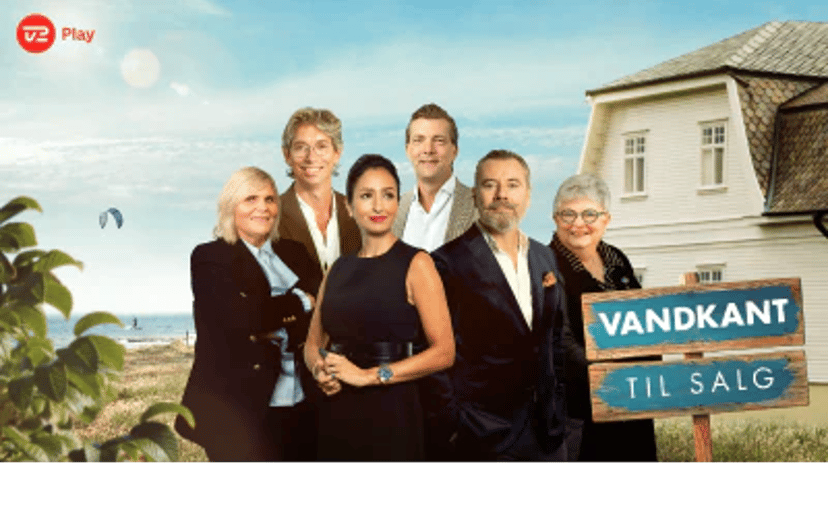 Fem personer står foran et hus ved vandet med et skilt med teksten “Vandkant til salg” og TV 2 Play-logo.