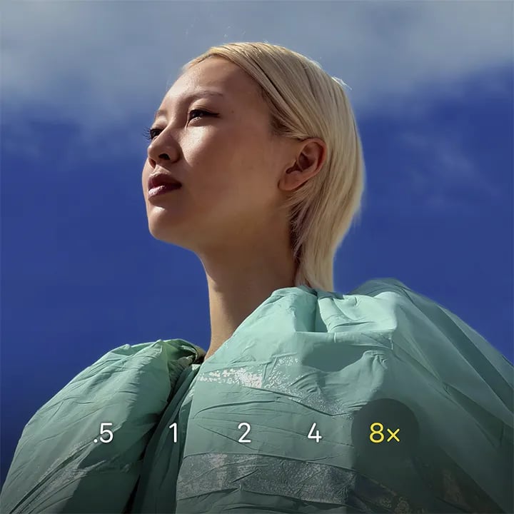 Portræt af en person med lyst hår i grøn jakke mod blå himmel, med zoommarkeringer fra 0,5x til 8x nederst.